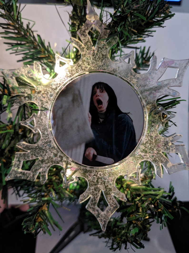 May include: A clear plastic snowflake ornament with a picture of a woman with dark hair and a white shirt, looking surprised and holding a knife. The ornament is decorated with glitter.