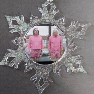 May include: A clear snowflake-shaped Christmas ornament with a glittery finish. The ornament features a photo of two people wearing pink shirts and shorts. The photo is set against a backdrop of a city street.