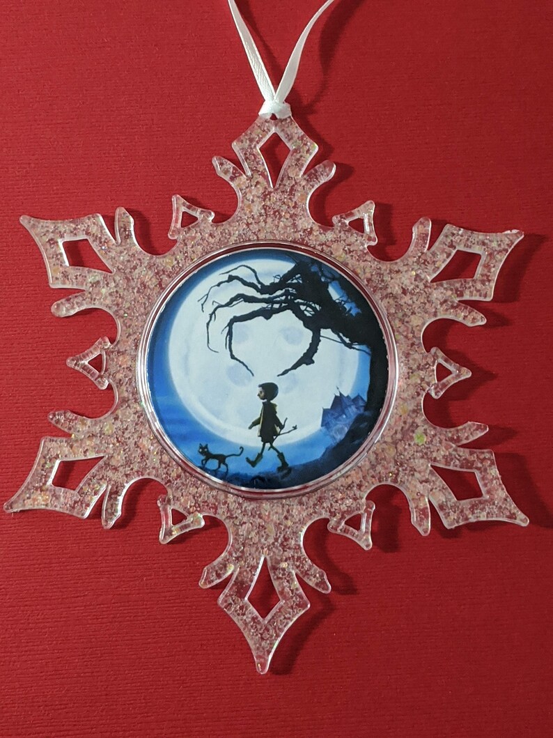May include: A clear snowflake-shaped ornament with a glittery finish. The ornament features a round image of a child walking with a cat in a moonlit forest.