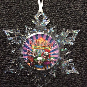 May include: A clear snowflake-shaped Christmas ornament with glitter. The ornament features a round, colorful image of four clowns with the text "Killer Klowns from Outer Space".