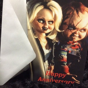 May include: A greeting card featuring a photo of the characters Chucky and Tiffany from the Child's Play horror film series. The card has the text "Happy Anniversary" in red.