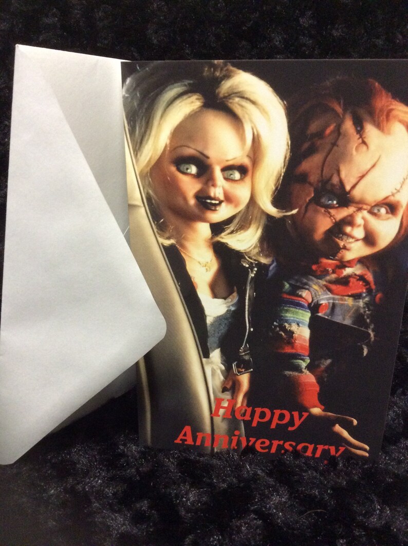 May include: A greeting card featuring a photo of the characters Chucky and Tiffany from the Child's Play horror film series. The card has the text "Happy Anniversary" in red.