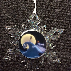 May include: A clear plastic snowflake ornament with a glittery finish. The ornament features a circular image of Jack Skellington and Sally from The Nightmare Before Christmas standing on a hill with a full moon in the background.