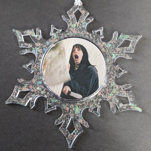 May include: A clear snowflake-shaped Christmas ornament with a glittery finish. The ornament features a round image of a woman with a shocked expression holding a knife.