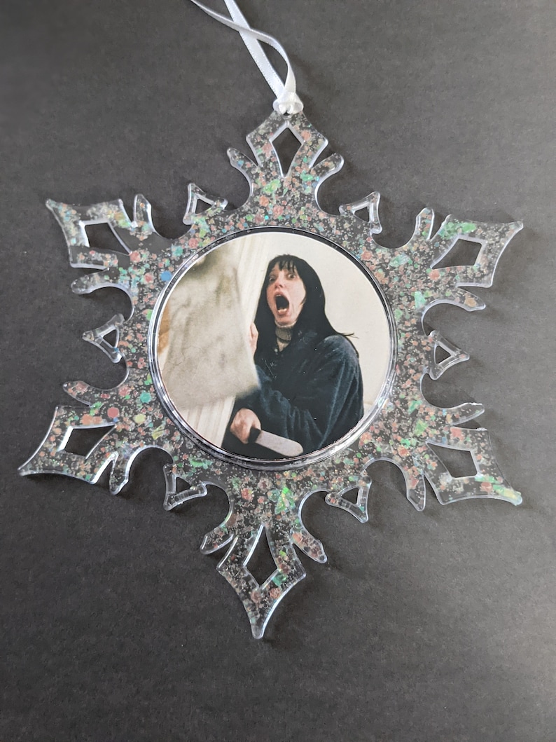 May include: A clear snowflake-shaped Christmas ornament with a glittery finish. The ornament features a round image of a woman with a shocked expression holding a knife.