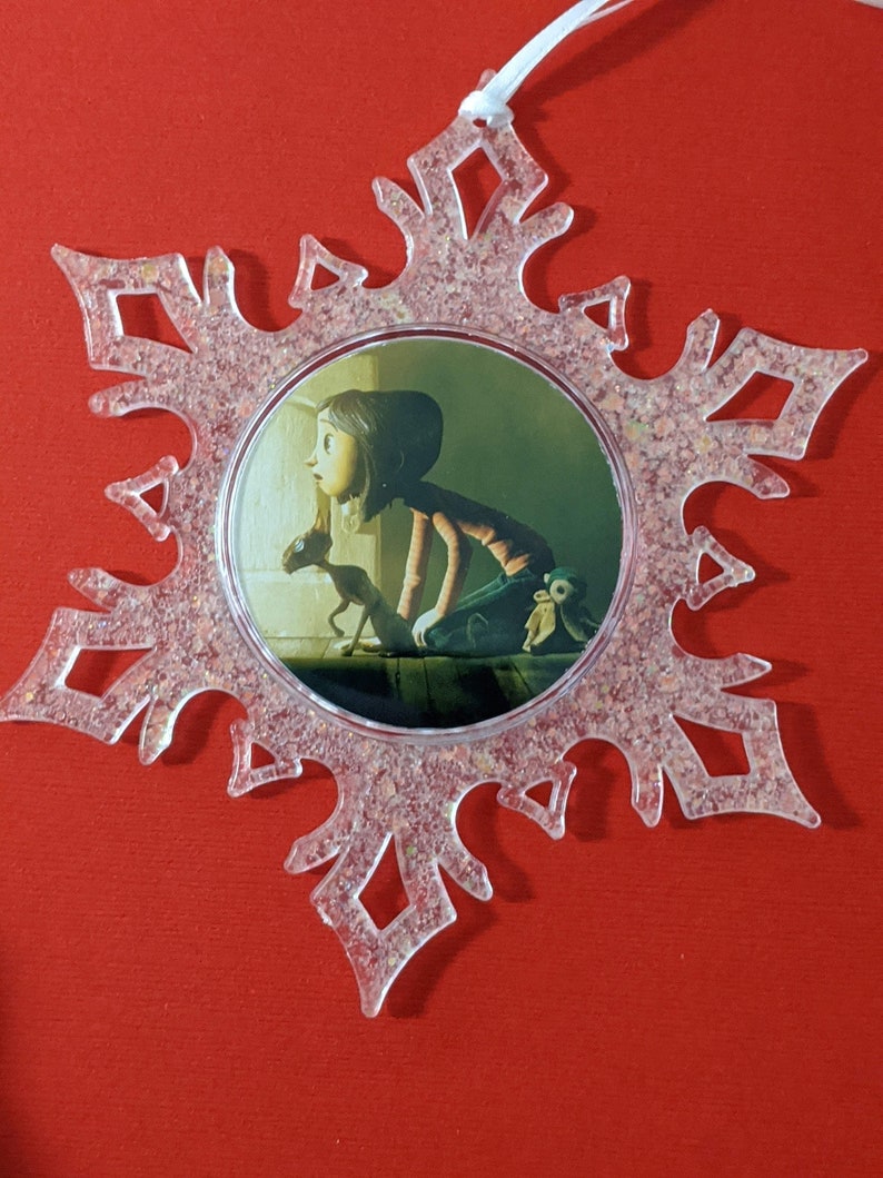 May include: A clear plastic snowflake ornament with a glittery finish. The ornament features a round image of a girl and a small creature in a dark room.