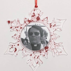 May include: A clear plastic snowflake ornament with a red ribbon. The ornament has red splatter paint and a black and white photo of a man wearing sunglasses and a clerical collar.