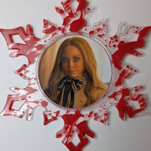 May include: A snowflake-shaped ornament with a portrait of a woman in the center. The clear plastic snowflake has red paint splatters and accents. A red ribbon is attached for hanging. The woman in the center has blonde hair and is wearing a black and white bow.