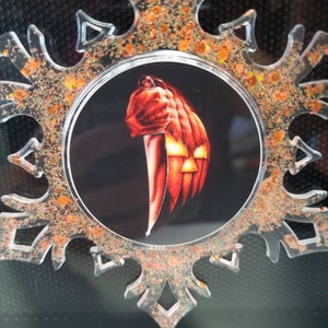 May include: A clear plastic snowflake ornament with orange glitter. The center of the snowflake features a round image of a hand holding a knife, with a pumpkin face in the background.