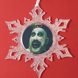 May include: A clear plastic snowflake ornament with a glittery pink finish. The ornament features a round image of a man with a painted face, wearing a red, white, and blue hat.