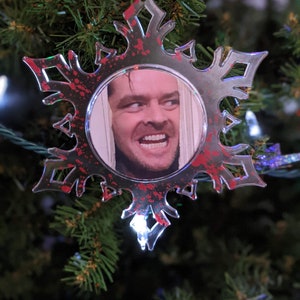 May include: A red and white snowflake bauble with a clear centre featuring a photo of a man with a sinister smile. The snowflake has a red splatter pattern.