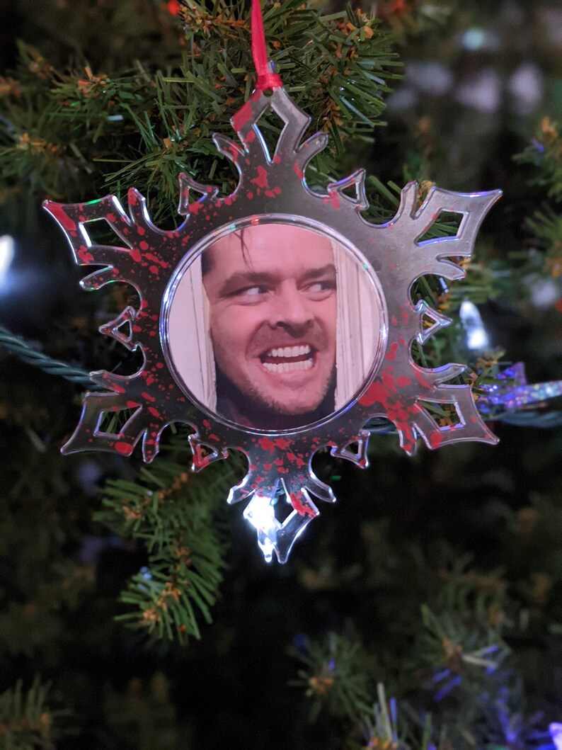 May include: A red and white snowflake bauble with a clear centre featuring a photo of a man with a sinister smile. The snowflake has a red splatter pattern.