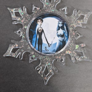 May include: A clear snowflake-shaped ornament with a glittery finish. The ornament features a circular image of Jack Skellington and Sally from the movie The Nightmare Before Christmas.