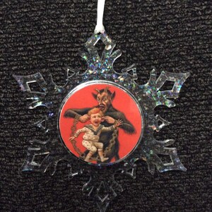 May include: A clear snowflake-shaped Christmas ornament with a red and black circular image of a devil holding a child by the ears. The ornament has a white string for hanging.