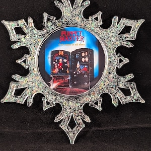 May include: A silver snowflake-shaped Christmas ornament with glitter. The ornament features a circular image of the movie "Puppet Master" with the text "Evil comes in all sizes".