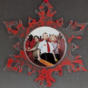 May include: A red and white snowflake Christmas ornament with a circular image of three people holding weapons. The image is surrounded by red splatter paint.