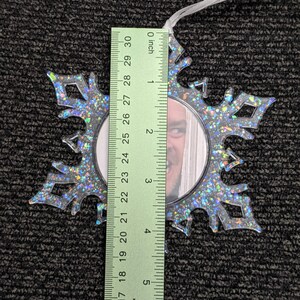 May include: A clear plastic snowflake ornament with a silver glitter finish. The ornament has a round opening in the centre that can be used to insert a photo. The ornament is approximately 7.6 cm in diameter.