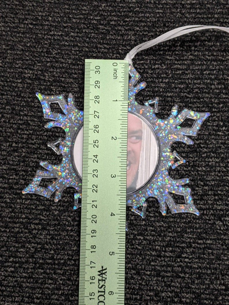 May include: A clear plastic snowflake ornament with a silver glitter finish. The ornament has a round opening in the centre that can be used to insert a photo. The ornament is approximately 7.6 cm in diameter.