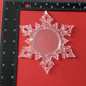 May include: A clear plastic snowflake ornament with a glitter finish and a round, clear window in the center. The ornament has a white string for hanging.