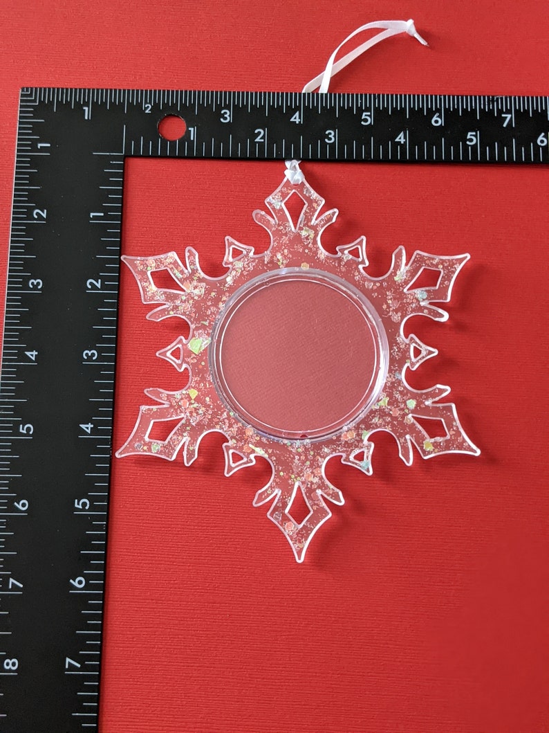 May include: A clear plastic snowflake ornament with a glitter finish and a round, clear window in the center. The ornament has a white string for hanging.