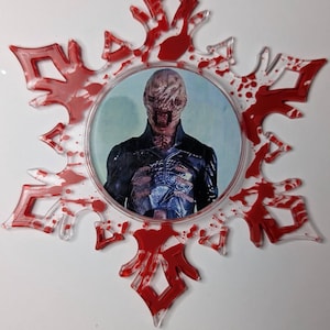 May include: A snowflake-shaped ornament with a central image of a character from a horror film. The ornament has a clear, transparent design with red accents resembling blood splatters. A red ribbon is attached for hanging.