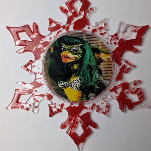 May include: A clear, snowflake-shaped ornament with red accents and a red ribbon for hanging. The center features a circular image of a Gremlin with green hair and a leopard print bikini. A unique holiday decoration.