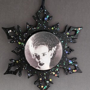 May include: Black snowflake ornament with iridescent glitter and a round image of the Bride of Frankenstein in the center.