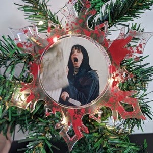 May include: A clear plastic snowflake ornament with red paint splatters. The ornament features a round photo of a woman with dark hair and a shocked expression holding a knife. The photo is surrounded by a silver ring.