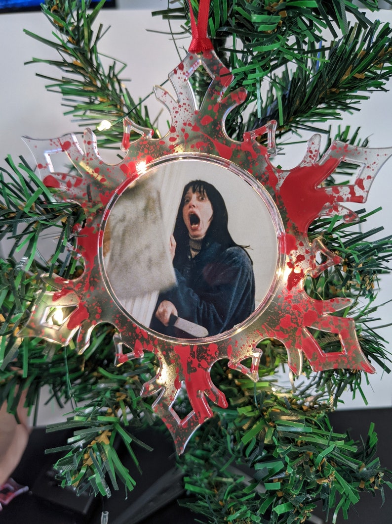 May include: A clear plastic snowflake ornament with red paint splatters. The ornament features a round photo of a woman with dark hair and a shocked expression holding a knife. The photo is surrounded by a silver ring.
