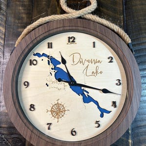 May include: Wooden wall clock with a light wood face, dark wood frame, and rope hanger. The clock face features a blue lake map, compass rose, and the words "Devonia Lake". Black clock hands and numerals.