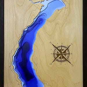 Peut inclure: Tableau en bois représentant une carte 3D du lac Comox, avec des nuances de bleu indiquant la profondeur. La carte est encadrée de noir et comprend une rose des vents. Le texte 'Comox Lake' est gravé sur le bois.