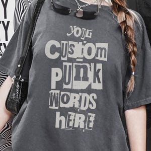 May include: Charcoal grey t-shirt featuring the text "YOUR CUSTOM PUNK WORDS HERE" in a distressed font. The shirt is paired with light trousers and a black handbag. A poster with the word "REALITY" is visible in the background.