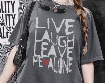 Può includere: Una maglietta grigia oversize con la scritta "Live Laugh Leave Me Alone" stampata in bianco con un cuore rosso che sostituisce la "e" di "me".