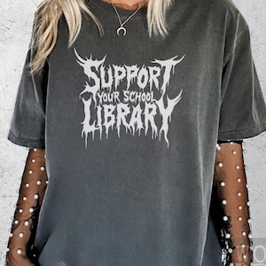 May include: A dark gray t-shirt with a white graphic that says "Support Your School Library" in a metal band font.