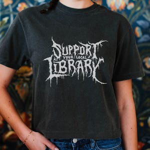 May include: Dark gray t-shirt with the text "SUPPORT YOUR LOCAL LIBRARY" in a white, stylized font. The shirt is short-sleeved and worn by a person. The background features a floral pattern.