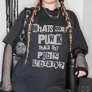 以下が含まれることがあります： 黒いオーバーサイズのTシャツに、「Whats More Punk Than The Public Library?」という文字が白でプリントされています。Tシャツは、長い三つ編みの髪と黒いチョーカーネックレスをした人が着用しています。