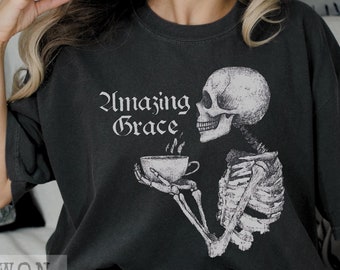 Pode incluir: Uma camiseta preta com um esqueleto segurando uma xícara de café fumegante. O texto "Amazing Grace" está impresso em branco na camiseta.