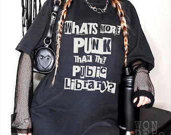May include: A black short-sleeved t-shirt featuring white text that reads "WHATS MORE PUNK THAN THE PUBLIC LIBRARY?" in a ransom-note style font. The shirt is worn over black fishnet sleeves and paired with a black heart-shaped crossbody bag.