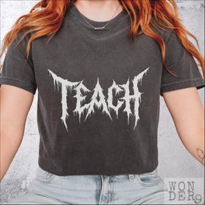 Könnte beinhalten: Dunkelgraues T-Shirt mit dem Wort "TEACH" in weißer, stacheliger, gotischer Schrift. Das kurzärmelige T-Shirt hat einen Rundhalsausschnitt. Der Text ist mittig auf der Vorderseite. Das T-Shirt ist aus weichem Material.