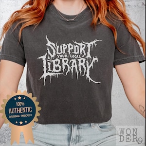 May include: A grey cropped t-shirt with a white graphic that reads "Support Your Local Library" in a metal band font. The shirt is being worn by a person with red hair. A gold star-shaped sticker with the text "100% Authentic Original Product" is in the lower left corner of the image.