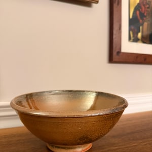 May include: A handmade ceramic bowl with a warm, earthy color palette. The bowl features a rounded shape with a slightly flared rim and a small, circular base. The interior has a glossy finish, while the exterior has a textured, matte appearance.