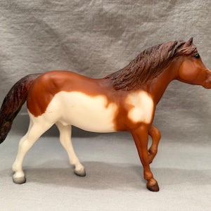 Può includere: Una statuetta di cavallo pinto marrone e bianco con una lunga criniera e coda. Il cavallo è in piedi su tutte e quattro le zampe con la testa girata di lato.
