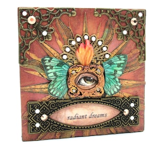 May include: A mixed media artwork featuring a large eye with a flame above it, surrounded by two green butterfly wings and a gold ornate frame. The background is a red and gold sunburst pattern with the text "radiant dreams" on a gold banner.