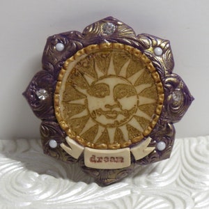 May include: A purple and gold sun with a face and the word "dream" on a banner. The sun is surrounded by a decorative border with white rhinestones.