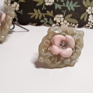 May include: A pair of earrings featuring a delicate pink flower design with a sparkly center, set in a light gray, textured base.