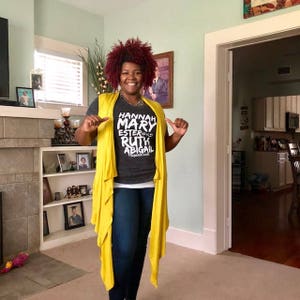 May include: A woman wearing a gray t-shirt with the text "HANNAH MARY ESTER RUTH ABIGAIL" printed on it. She is also wearing a yellow cardigan and blue jeans.