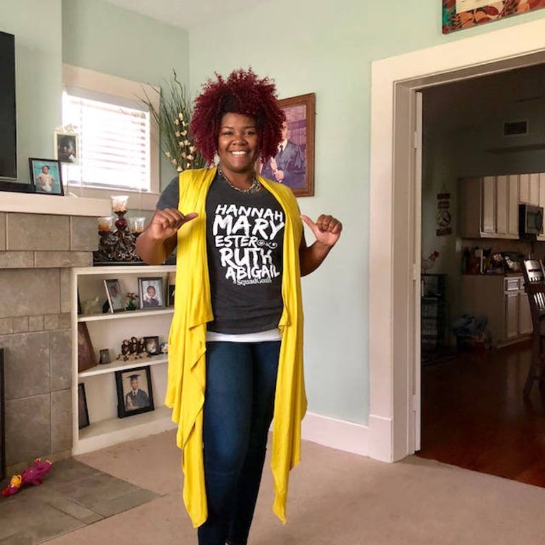 May include: A woman wearing a gray t-shirt with the text "HANNAH MARY ESTER RUTH ABIGAIL" printed on it. She is also wearing a yellow cardigan and blue jeans.