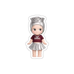 May include: A doll figurine with a silver rose headpiece, a maroon t-shirt with the number "1986", and a silver pleated skirt. The doll has a necklace and is standing with its arms outstretched.