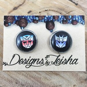 May include: A pair of round, black stud earrings with the Autobot and Decepticon symbols from Transformers. The earrings are set in silver-coloured metal and displayed on a tan card with the text "Designs by Leisha".