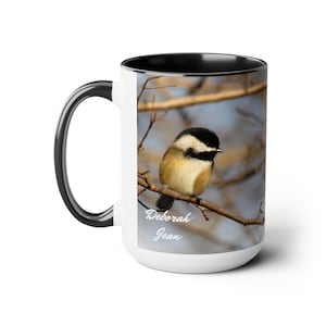 May include: White ceramic coffee mug with a black rim and handle. The mug features a photograph of a black-capped chickadee perched on a branch with a blurred background of trees and sky. The text "Deborah Jean" is printed on the mug.